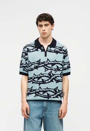 Mladić u svijetloplavoj polo majici kratkih rukava s crnim apstraktnim uzorkom i plavim trapericama, stoji ispred jednobojne bijele pozadine.