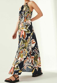 Robe maxi florale avec un fond marine, présentant un motif de fleurs colorées, un col halter et des fentes sur les côtés. Portée avec des sandales noires à lanières.