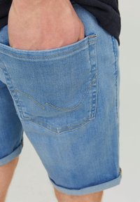 Person wearing light blue denim shorts with hand resting inside the back pocket against a plain background.