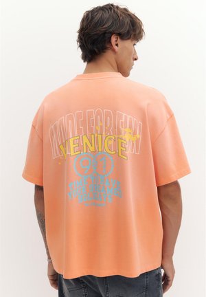 Mladý muž s krátkými vlasy nosí broskvové tričko s nápisy „Made for Fun“, „Venice 90“ a „Time to have your dreamed holidays“ na zádech.