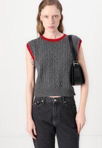 Pull en tricot sans manches gris avec un col et des accents de bras côtelés rouges, arborant un motif en câble texturé, associé à un jean noir taille haute.