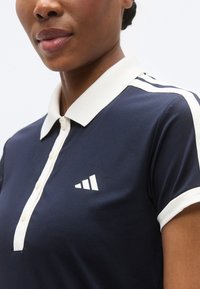 Polo bleu marine avec col blanc et patte de boutonnage. Comprend des bandes blanches sur les côtés et un petit logo blanc sur le côté gauche de la poitrine.