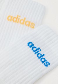 Λευκές ριγέ κάλτσες με το λογότυπο "adidas" σε κίτρινο και μπλε. Το σχέδιο περιλαμβάνει αντιθετικά χρώματα σε κάθε κάλτσα. Μαλακή υφή.