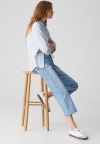Lichtblauw button-up shirt, relax-fit lichtblauwe jeans en witte sneakers met bruine zolen, zittend op een houten kruk tegen een neutrale achtergrond.