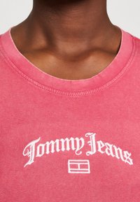 Červené bavlněné tričko s kulatým výstřihem, na kterém je bílý text "Tommy Jeans" a pod ním obdélníkové logo. Měkká textura, ležérní design.