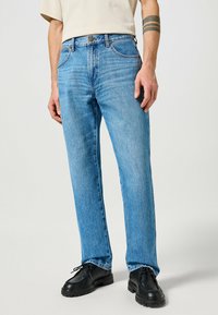 Jean droit en denim bleu clair avec une texture délavée, design à cinq poches et fermeture à bouton en métal. Porté avec des chaussures noires.