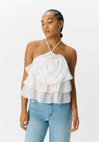 Witte gehaakte haltertop met gelaagd bloemblaadjesontwerp, voorzien van een textuurstof en een ruitpatroon. Gekleed met lichtblauwe denimjeans.