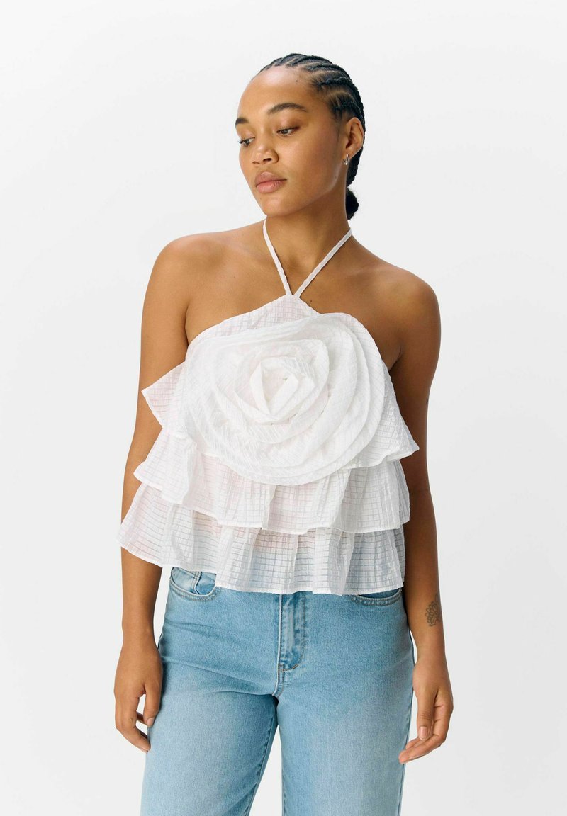 Witte gehaakte haltertop met gelaagd bloemblaadjesontwerp, voorzien van een textuurstof en een ruitpatroon. Gekleed met lichtblauwe denimjeans.