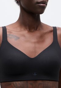 Κοντινό πλάνο ενός ατόμου που φοράει μαύρο αθλητικό σουτιέν με μικρό λογότυπο στο μπροστινό μέρος και ορατό τατουάζ στο πάνω μέρος του στήθους και κάτω από το σουτιέν.