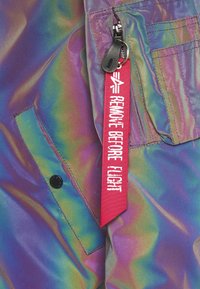 Irydescencyjny materiał w fioletowych i zielonych odcieniach o matowej teksturze. Posiada czarny guzik i czerwoną metkę z napisem „ZDJĄĆ PRZED LOTEM.”