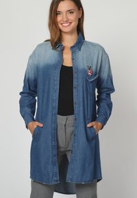 Camisa de mezclilla con un color azul degradado, de mangas largas, cierre delantero con botones, dos bolsillos laterales y un parche de símbolo de paz en el pecho.