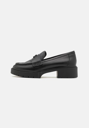 Mocassin en cuir noir avec dessus texturé, pompon décoratif, talon épais et semelle robuste, présenté de profil sur fond blanc.
