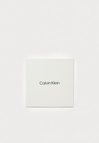 Biely štvorcový box s textom "Calvin Klein" zobrazeným tučným čiernym písmom uprostred prednej strany. Hladký matný povrch bez ďalších dizajnov.