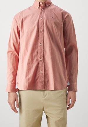 Homme portant une chemise rose à manches longues boutonnée avec un pantalon beige, debout devant un fond clair uni.