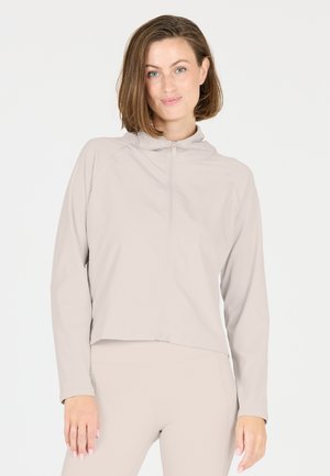 Femme aux cheveux courts et bruns portant un haut beige à manches longues zippé et un pantalon assorti, debout devant un fond blanc.