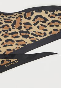 Foulard en soie avec un motif léopard beige et noir, bordé de noir. Logo Coach en or à une extrémité. Forme rectangulaire, texture lisse.