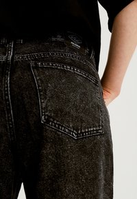 Nära bild av en person som bär svarta acid-wash-jeans med en synlig bakficka och en pull&bear-märkesetikett, handen vilande på höften.