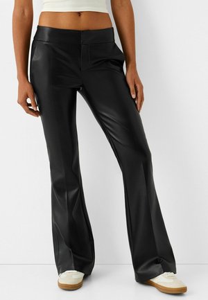 Kobieta ubrana w wysokie, czarne spodnie dzwony z imitacji skóry i białe trampki, stojąca na tle jednolitego białego tła.