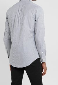 Mężczyzna ubrany w dopasowaną białą koszulę w cienkie pionowe czarne paski, w zestawieniu z czarnymi spodniami, widoczny z tyłu.