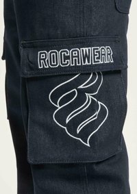 Tumman siniset cargo-housut, joissa on läppäpussi "ROCAWEAR"-logolla ja valkoisella brodeeratulla spiraalikuviolla sivussa. Raskas rakenne, monipuolinen taskusuunnittelu.