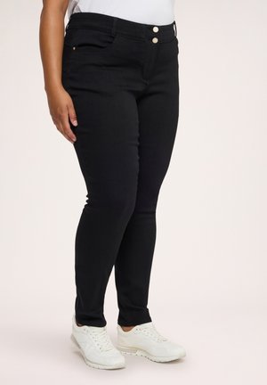 Person iført tætsiddende sorte jeans med dobbelt knaplukning og hvide sneakers, stående mod en ensfarvet lys baggrund.