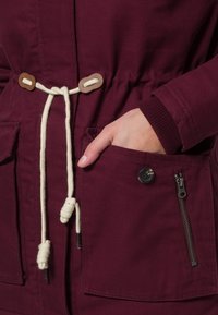 Burgundy jacka med krämfärgad dragsko och träknopp, elastiska ärmslut, framfickor och en zip-ficka på sidan. Hållbart tyg.