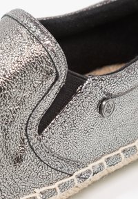 Chaussure argentée en tissu texturé à enfiler, avec un panneau élastique noir sur le côté, un détail de logo métallique rond et une couture de semelle tissée.