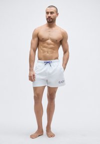 Fehér úszónadrág kék húzózsinórral, sötétkék "EA7" logóval. Az egyszerű és könnyű kialakítás strandviseletre ideális.