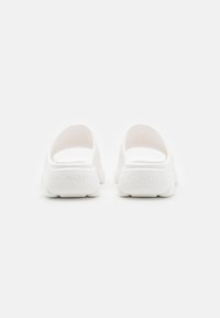 Zapatillas blancas sin cordones con una textura suave, forma redondeada y un pequeño logo en la parte trasera. El diseño de talón abierto mejora la transpirabilidad.