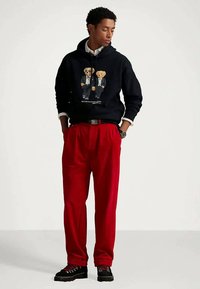 Νεαρός άνδρας με μαύρη κουκούλα που έχει γραφικά με δύο αρκουδάκια, κόκκινο παντελόνι, μαύρες μπότες και λευκό πουκάμισο, στέκεται με τα χέρια στις τσέπες.