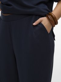 Persoon draagt donkerblauwe broek met hand in de zak en twee grote houten armbanden om de pols tegen een witte achtergrond.