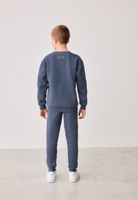 Blauer Pullover und passende Hose aus weichem Stoff, mit einem Etikett auf der Rückseite und weißen Turnschuhen. Glatte Textur und entspannte Passform.