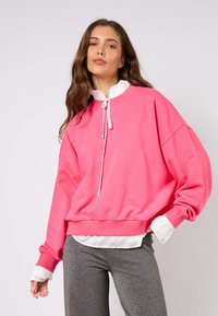 Nadrozmerná ružová mikina s kapucňou na sťahovanie, vrstvená cez bielu košeľu s volánmi. Kombinovaná s trblietavými sivými nohavicami.