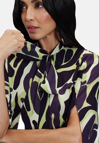 Blusa a maniche corte con un motivo astratto viola e verde lime, colletto a fiocco e tessuto testurizzato.