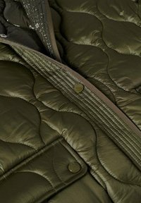Olivově zelená prošívaná bunda s lesklou texturou, kontrastním lemováním, patentkami a odhaleným zipem na přední straně.