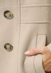 Beige wollen jas met een gestructureerde afwerking, voorzien van twee grote, gemusterde knopen en een zijzak. Zachte stof met getailleerde lijnen.