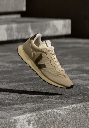 Beige en bruine sneaker met "V"-logo, zwevend boven een gestructureerd grijs betonnen oppervlak en trappen op de achtergrond.