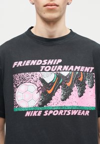 Κοντινό πλάνο ενός ατόμου που φοράει μαύρο μπλουζάκι με ροζ, πράσινα και πορτοκαλί γραφικά από παπούτσια ποδοσφαίρου που κλωτσούν μια μπάλα και το κείμενο «Friendship Tournament Nike Sportswear».