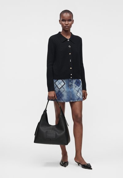Femme portant un cardigan noir boutonné et une mini-jupe en patchwork denim, tenant un grand sac à main noir avec des talons noirs pointus.