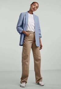 Lys blå blazer over en hvit t-skjorte, kombinert med beige vide bukser og hvite joggesko. Glatt stoff med en avslappet passform.