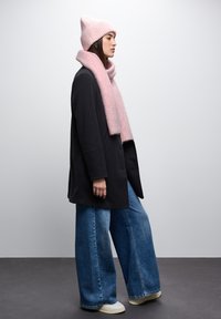 Cappotto oversize nero, berretto a coste rosa chiaro, sciarpa abbinata, jeans a gamba larga blu e sneakers crema. Abbigliamento invernale semplice e comodo.