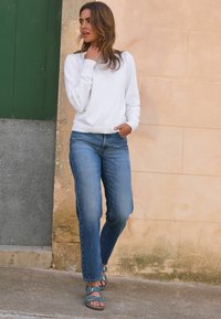 Witte lange mouwen trui met geribbelde manchetten, blauwe rechte jeans en blauwe sandalen met gespen. De achtergrond toont een gecementeerde stenen muur.