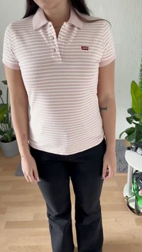 Donna che indossa una polo Levi's a righe rosa chiaro e bianco e pantaloni neri, in piedi all'interno vicino a piante e a un tavolo bianco.