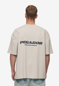 Vaalea beige suuri T-paita lyhyillä hihoilla, jossa on musta painettu logo "PEGADOR" selkäpuolella. Pehmeää puuvillamateriaalia.