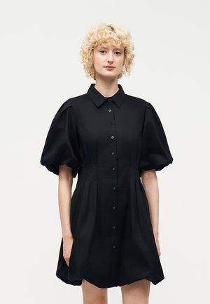 Kvinde med krøllet blondt hår iført en sort kjole med puffede korte ærmer og en knaplukning foran, stående mod en ensfarvet baggrund.