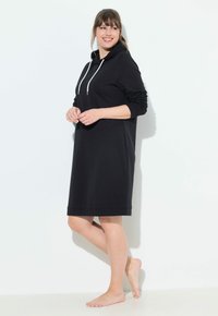 Donna sorridente a piedi nudi che indossa un vestito con cappuccio nero a maniche lunghe, in piedi di lato su uno sfondo bianco semplice.