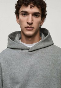 Felpa grigia con una texture morbida, cappuccio ampio e tasca a marsupio. Indossata sopra una t-shirt bianca a girocollo per creare un effetto di stratificazione.
