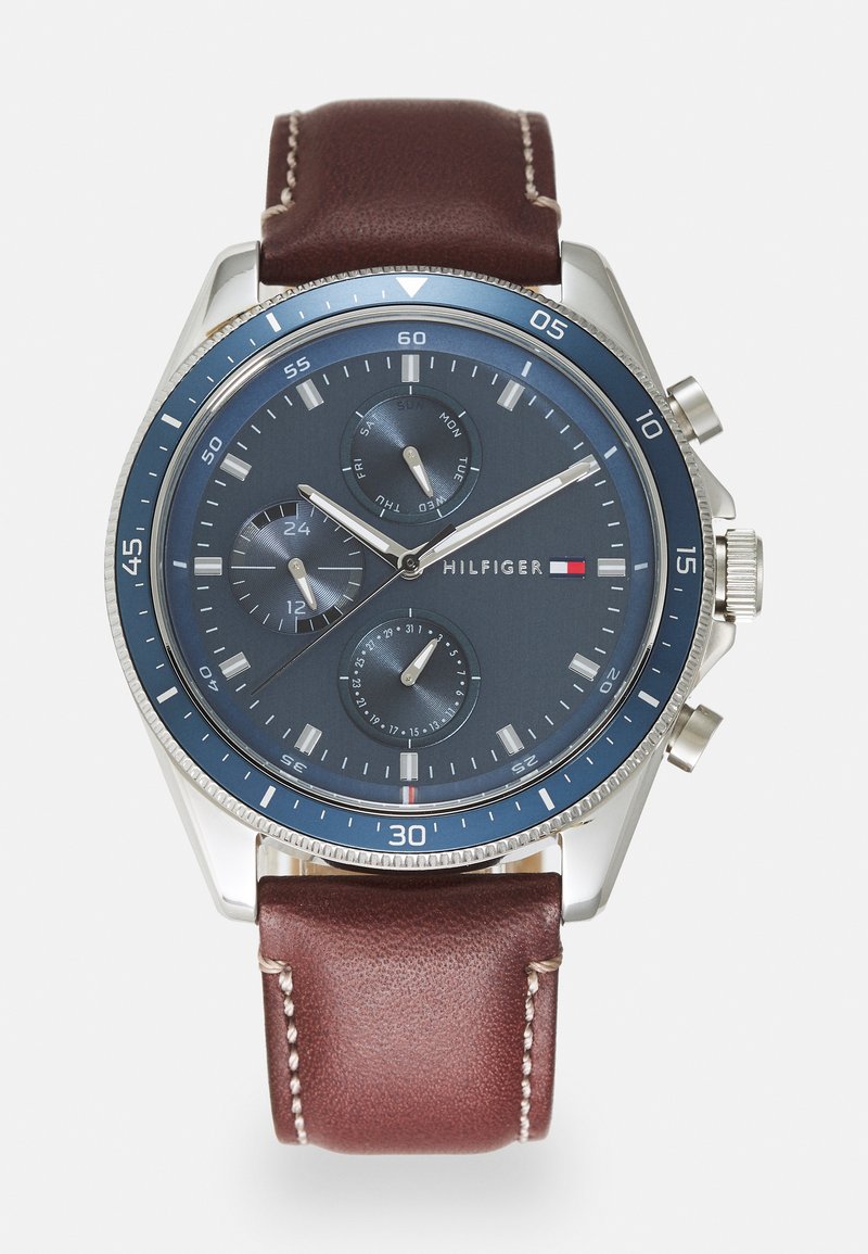 Reloj de acero inoxidable con bisel azul, esfera azul oscura y acentos plateados. Correa de cuero marrón con costuras blancas y hardware pulido.