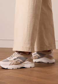 Sneakers beige con pannelli in rete e suede, suola intermedia bianca imbottita e lacci rotondi. Abbinati a pantaloni larghi di colore crema.