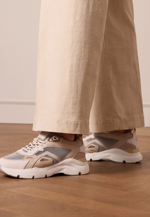 Sneakers beige con pannelli in rete e suede, suola intermedia bianca imbottita e lacci rotondi. Abbinati a pantaloni larghi di colore crema.